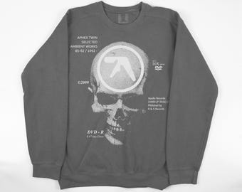 May include: Gray sweatshirt featuring a white skull graphic with the Aphex Twin logo and text. The text includes "APHEX TWIN SELECTED AMBIENT WORKS 85-92/1992-" and "DVD-R". The sweatshirt has a crew neck and long sleeves.