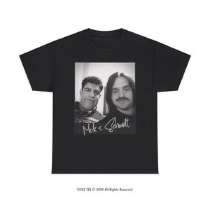 Op de afbeelding: Zwart-witfoto van twee mannen, een met een baard en snor, de ander met een geschoren gezicht. De foto is gedrukt op een zwart T-shirt. De tekst "Nick < Schmidt" is in wit onder de foto geschreven.