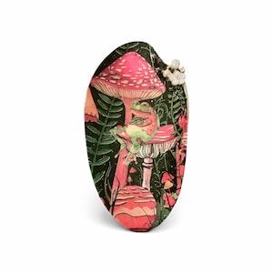 May include: An oval-shaped pendant featuring a whimsical illustration of a frog on a mushroom. The design includes pink and red mushrooms, green foliage, and a dark background. A small white flower is attached to the top of the pendant.