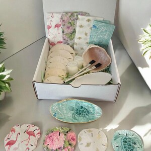 Puede incluir: Una caja blanca llena de art&iacute;culos con tem&aacute;tica playera, incluyendo conchas marinas, d&oacute;lares de arena, servilletas y velas. La caja est&aacute; abierta y los art&iacute;culos son visibles. Tambi&eacute;n hay cinco posavasos con forma de d&oacute;lar de arena con diferentes dise&ntilde;os, incluyendo flamencos rosas, patrones florales y una tortuga.
