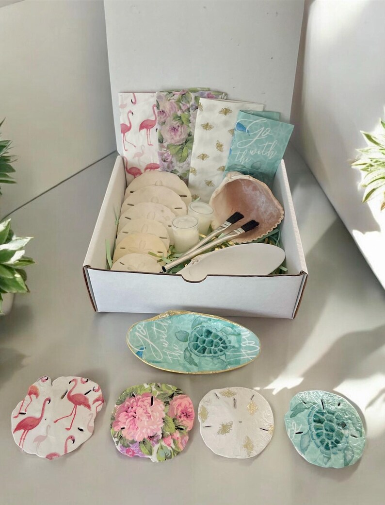 Puede incluir: Una caja blanca llena de art&iacute;culos con tem&aacute;tica playera, incluyendo conchas marinas, d&oacute;lares de arena, servilletas y velas. La caja est&aacute; abierta y los art&iacute;culos son visibles. Tambi&eacute;n hay cinco posavasos con forma de d&oacute;lar de arena con diferentes dise&ntilde;os, incluyendo flamencos rosas, patrones florales y una tortuga.
