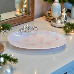 May include: Oval-shaped decorative serving dish with a pink and white marble design. The dish is adorned with a cluster of faux pearls and crystals. The dish is approximately 12 inches long.