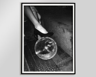 Puede incluir: Fotografía en blanco y negro de una bola de discoteca sobre un suelo de madera. Se ven las piernas y los pies de una persona, con un pie calzado con un zapato de tacón negro. La bola de discoteca refleja la luz sobre el suelo y una superficie cercana.