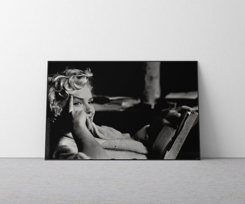 May include: A black and white framed wall art print featuring a portrait of Marilyn Monroe with her signature blonde curls, resting her chin on her hand while holding an open book, captured in a candid moment with high contrast lighting.