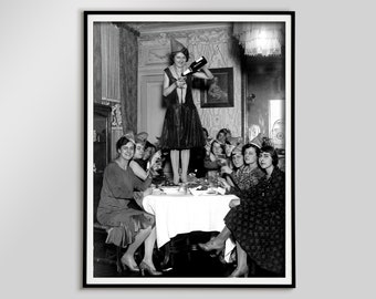 May include: A monochrome framed print depicting a vintage party. A woman in a party hat pours champagne into glasses, while others seated around a table raise their glasses. The image captures a celebratory moment.