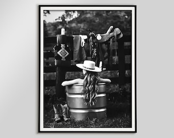 May include: Black and white photograph of a person in a metal tub, wearing a white hat, with long hair. Cowboy boots are at the base of the tub. A saddle and other items hang on a wooden fence in the background.