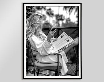 May include: Black and white framed print of a person sitting in a chair on a balcony, holding a glass of red wine and reading a newspaper. The person is wearing a white robe. Palm trees are visible in the background.