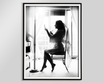 Può includere: Fotografia in bianco e nero incorniciata di una persona in silhouette, seduta e che si trucca. La persona indossa un accappatoio e un velo, con una finestra e delle tende sullo sfondo. È visibile una lampada da terra.
