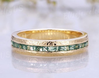 Puede incluir: Un anillo dorado con textura martillada y una banda de pequeñas piedras preciosas verdes de corte cuadrado. El anillo está centrado, con un fondo suave y borroso de flores blancas.