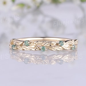 Pode incluir: Um anel de ouro com um design de folhas e pequenas pedras preciosas redondas verde claro. O anel está sobre um fundo desfocado de flores brancas.