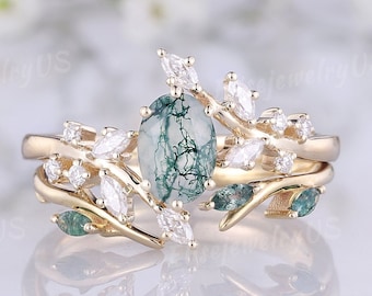 May include: A gold ring set featuring an oval-cut moss agate gemstone, surrounded by marquise-cut diamonds and leaf accents. The ring has a nature-inspired design with a delicate, botanical aesthetic. The ring is set on a reflective surface.