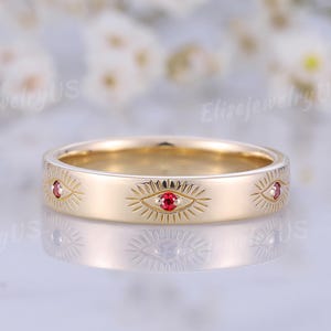 Peut inclure: Une bague anneau en or ornée d'une série de motifs d'yeux gravés, chacun centré par une petite pierre précieuse ronde d'un rouge éclatant. La surface métallique est lisse et polie, présentant un motif répétitif de lignes rayonnantes.