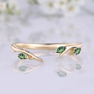 Puede incluir: Un anillo dorado con un diseño en forma de rama. El anillo presenta tres piedras verdes con corte marquesa, que se asemejan a hojas. El fondo es suave y borroso.