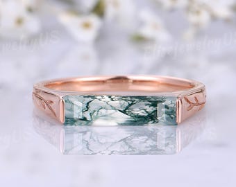 Puede incluir: Un anillo de oro rosa con una piedra rectangular de ágata musgosa. La piedra es de color verde claro con vetas verde oscuro. El anillo tiene un diseño de hojas en los laterales.