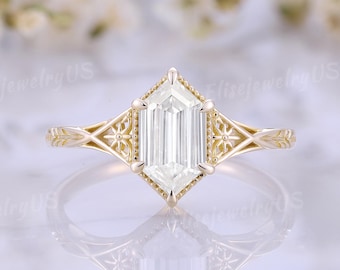 以下が含まれることがあります： 大きなエメラルドカットの白い宝石が付いたゴールドの婚約指輪。指輪は、バンドと中央の石の周りに複雑なフィリグリーのディテールがあり、幾何学的なデザインになっています。