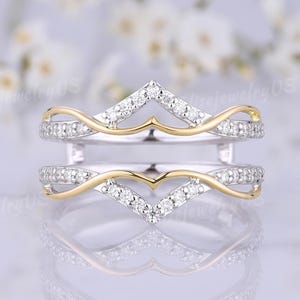 May include: A two-piece ring guard featuring a chevron-shaped design with white metal bands encrusted with small clear stones and accented by thin, smooth yellow gold-toned metal curves that frame the center space for a solitaire ring.