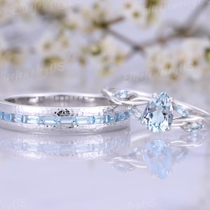 May include: Two matching wedding rings. One ring features a band with a hammered texture and light blue rectangular stones. The other ring has a pear-shaped light blue stone and smaller accent stones. The rings are silver in color.