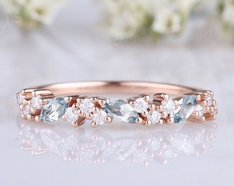 May include: A delicate rose gold band with a row of alternating aquamarine and white gemstones. The ring is set on a white background.