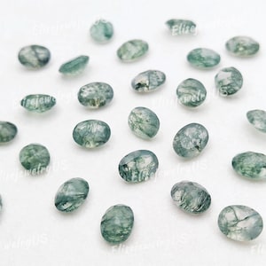 Puede incluir: Una colección de piedras preciosas de ágata musgosa de forma ovalada, cada una con un patrón único de inclusiones verdes en una base translúcida. Las piedras están dispersas sobre una superficie blanca.