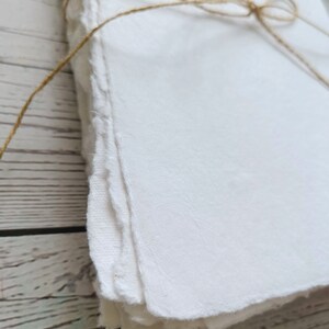 May include: A stack of handmade white paper with rough, deckled edges, tied with natural twine. The paper has a textured surface and appears to be thick. The background is a light-colored wooden surface.