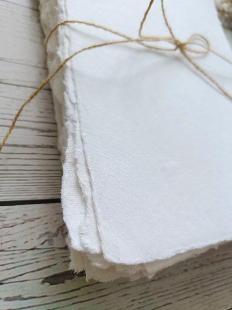 May include: A stack of handmade white paper with rough, deckled edges, tied with natural twine. The paper has a textured surface and appears to be thick. The background is a light-colored wooden surface.