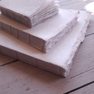 May include: Handmade white paper journals with deckled edges, stacked in a tiered arrangement. The spines show visible stitching. The paper has a textured, slightly uneven surface, giving a natural, artisanal feel. The journals are of varying sizes, creating a visually appealing display.