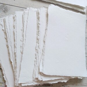 May include: A stack of handmade, white paper sheets with rough, deckled edges. The paper has a textured surface and appears to be suitable for art or calligraphy. The paper is stacked on a light-colored wooden surface.