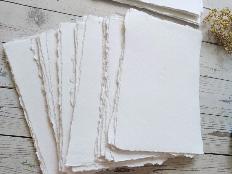 May include: A stack of handmade, white paper sheets with rough, deckled edges. The paper has a textured surface and appears to be suitable for art or calligraphy. The paper is stacked on a light-colored wooden surface.