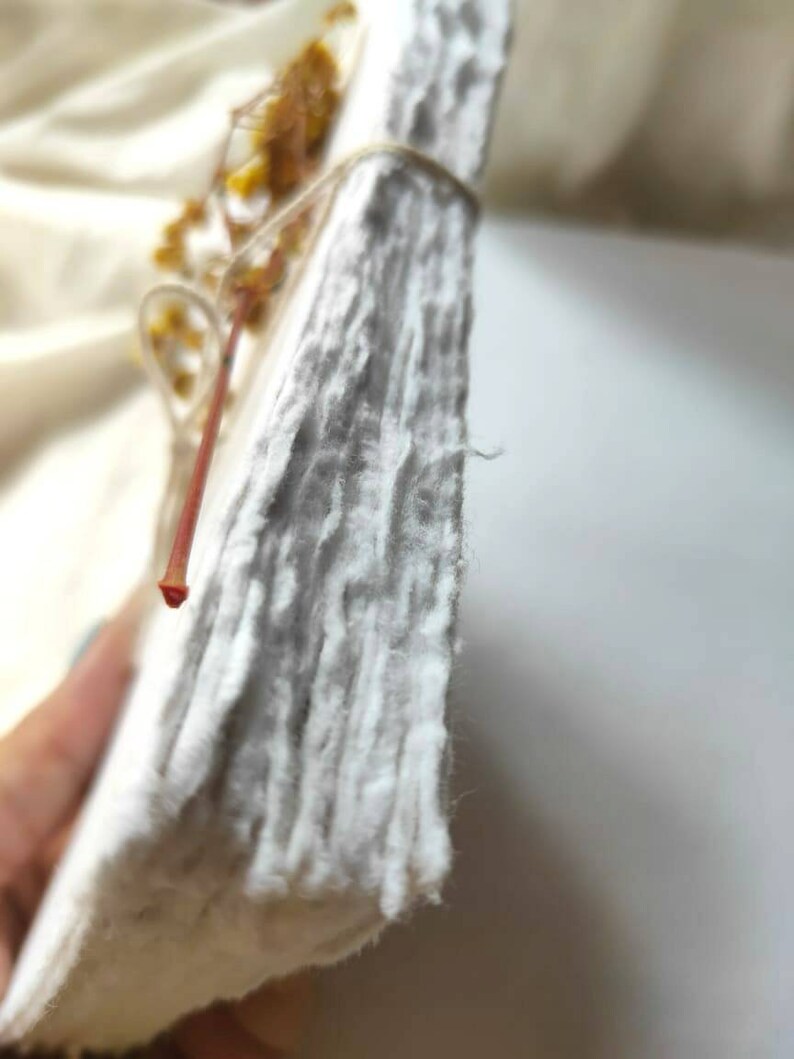 May include: A stack of handmade, textured white paper, bound with twine and adorned with dried yellow flowers. The paper has a rough, uneven surface, suggesting a natural, artisanal quality. The overall aesthetic is rustic and organic.