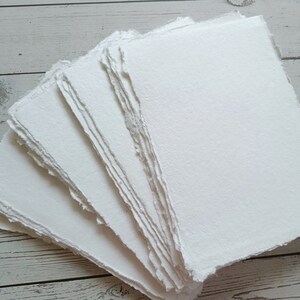 May include: A stack of handmade, white paper sheets with rough, deckled edges. The paper has a textured surface and appears to be thick and high-quality. The sheets are arranged in a fan shape, showcasing the texture and edges.