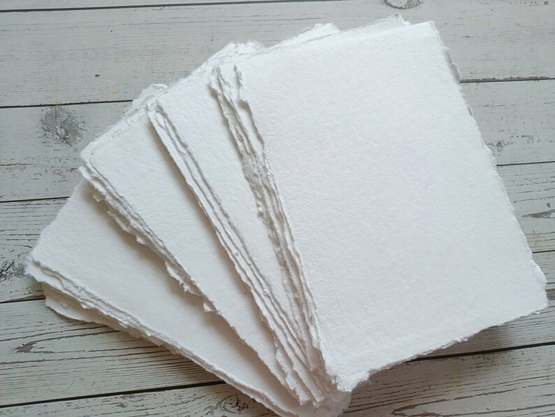 May include: A stack of handmade, white paper sheets with rough, deckled edges. The paper has a textured surface and appears to be thick and high-quality. The sheets are arranged in a fan shape, showcasing the texture and edges.