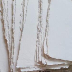 May include: A stack of handmade, off-white paper sheets with deckled edges. The paper has a slightly textured surface and a soft, natural appearance. The edges are uneven, giving the paper a rustic, artisanal feel.