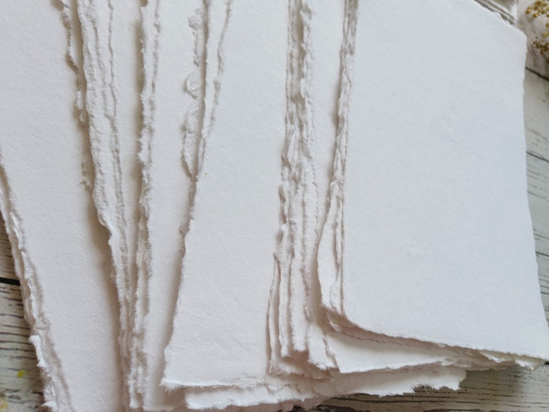 May include: A stack of handmade, off-white paper sheets with deckled edges. The paper has a slightly textured surface and a soft, natural appearance. The edges are uneven, giving the paper a rustic, artisanal feel.