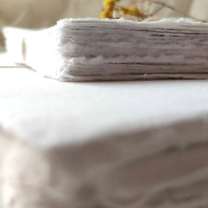 May include: A stack of handmade, deckle-edged paper, with a rough, textured appearance. The paper is a soft white color, and the edges are uneven, giving it a rustic feel. A few sprigs of yellow flowers are visible in the background.