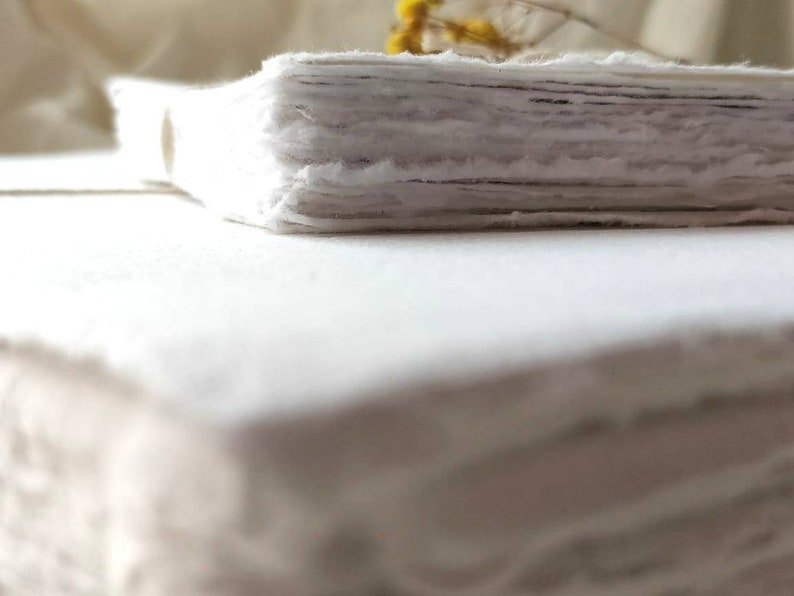 May include: A stack of handmade, deckle-edged paper, with a rough, textured appearance. The paper is a soft white color, and the edges are uneven, giving it a rustic feel. A few sprigs of yellow flowers are visible in the background.