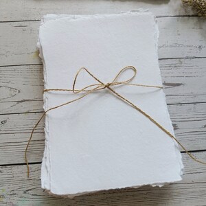 May include: A stack of handmade white paper with deckled edges, tied with twine. The paper has a textured surface and is arranged on a light-colored wooden surface. Additional stacks of paper are visible in the background.