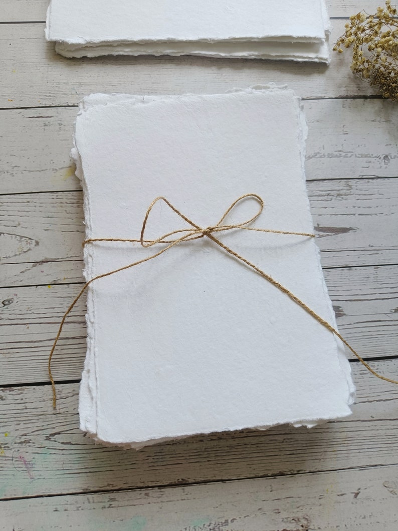 May include: A stack of handmade white paper with deckled edges, tied with twine. The paper has a textured surface and is arranged on a light-colored wooden surface. Additional stacks of paper are visible in the background.