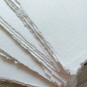 May include: A stack of handmade, off-white paper sheets with deckled edges. The paper has a slightly textured surface and a soft, natural appearance. Ideal for art projects, calligraphy, or journaling.