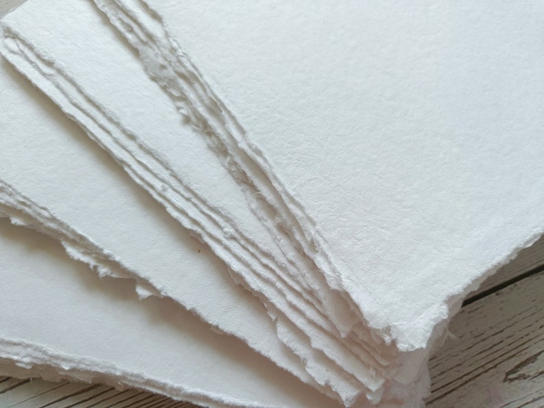 May include: A stack of handmade, off-white paper sheets with deckled edges. The paper has a slightly textured surface and a soft, natural appearance. Ideal for art projects, calligraphy, or journaling.