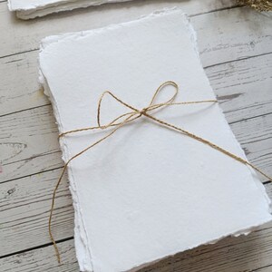 May include: A stack of handmade white paper with deckled edges, tied with twine. The paper has a textured surface and is arranged on a light wood background. Dried flowers are in the background.