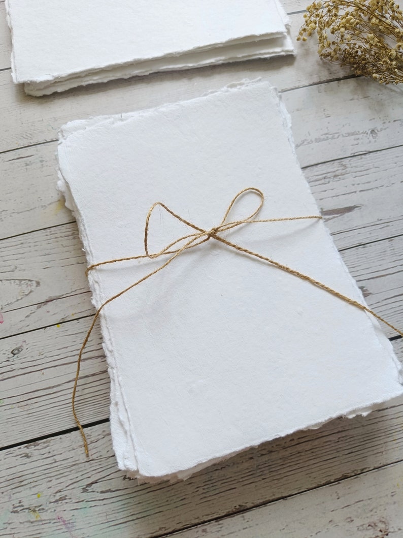 May include: A stack of handmade white paper with deckled edges, tied with twine. The paper has a textured surface and is arranged on a light wood background. Dried flowers are in the background.