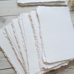 May include: A stack of handmade, white paper sheets with deckled edges. The paper has a slightly textured surface and is arranged in a fan shape, with additional stacks in the background. The paper is likely for art or calligraphy.