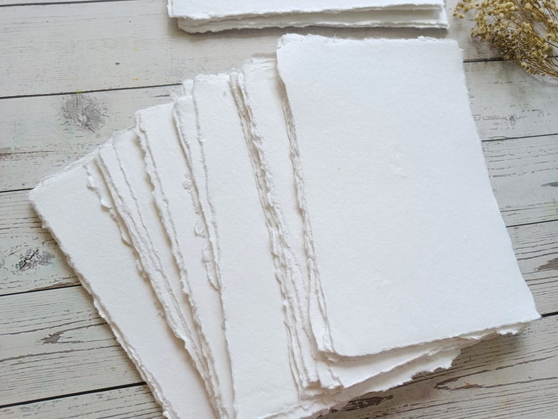 May include: A stack of handmade, white paper sheets with deckled edges. The paper has a slightly textured surface and is arranged in a fan shape, with additional stacks in the background. The paper is likely for art or calligraphy.