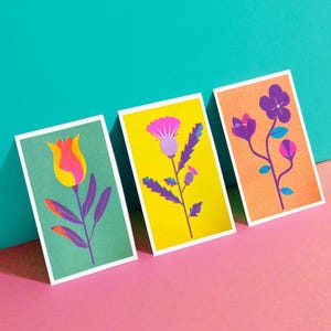 Può includere: Tre stampe artistiche rettangolari con illustrazioni floreali. La prima mostra un tulipano giallo e rosa su sfondo verde. La seconda presenta un cardo rosa su sfondo giallo. La terza raffigura fiori viola su uno sfondo di colore arancione.