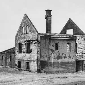 Peut inclure: Une photographie en noir et blanc d'un bâtiment délabré et abandonné avec une cheminée. Le bâtiment est en béton et possède plusieurs fenêtres et portes. Le bâtiment est entouré d'un champ d'herbe.