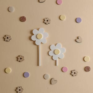 May include: A collection of decorative wooden shapes and flowers in various sizes and colors. The image features two large white daisy-shaped objects with yellow centers, along with smaller wooden flowers, hearts, and circular shapes in pastel colors and glitter.