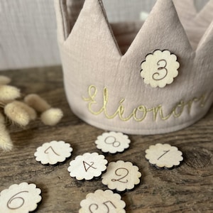 Könnte beinhalten: Eine hellbeige Stoffkrone mit goldgesticktem Namen und einer Holznummer 3. Mehrere blumenförmige Holzmarken mit den Zahlen 1-8 sind um die Krone verstreut. Die Krone ist für einen Kindergeburtstag bestimmt.