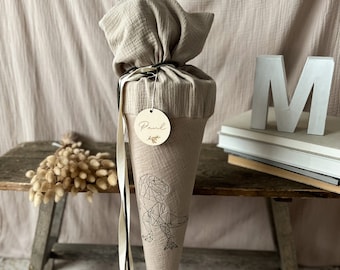 May include: A beige fabric cone with a geometric dinosaur design and a wooden tag with the name "Paul" on it. The cone is decorated with black and white ribbon and is sitting on a wooden table with a stack of books and a white letter "M".