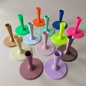 Könnte beinhalten: Eine Sammlung farbenfroher, moderner Kerzenhalter. Jeder Halter hat einen kreisförmigen Sockel und eine schlanke, vasenartige Spitze. Die Halter sind in verschiedenen Farben erhältlich, darunter Grün, Orange, Rosa und Blau. Die Sockel haben einen Durchmesser von etwa 10 cm.
