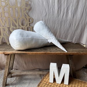 Könnte beinhalten: Zwei weiße, tropfenförmige Zierkissen liegen auf einer rustikalen Holzbank. Ein großer Alphabet-Ausschnitt aus Holz und ein weißer Buchstabe "M" befinden sich in der Nähe. Der Hintergrund zeigt einen strukturierten, hellfarbigen Stoff.